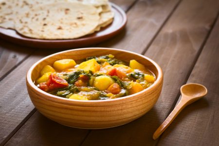 Wooden bowl of pumpkin, mangold, potato and tomato curry dish with homemade chapati flatbread in the back photographed with natural light (Selective Focus, Focus in the middle of the curry dish)
